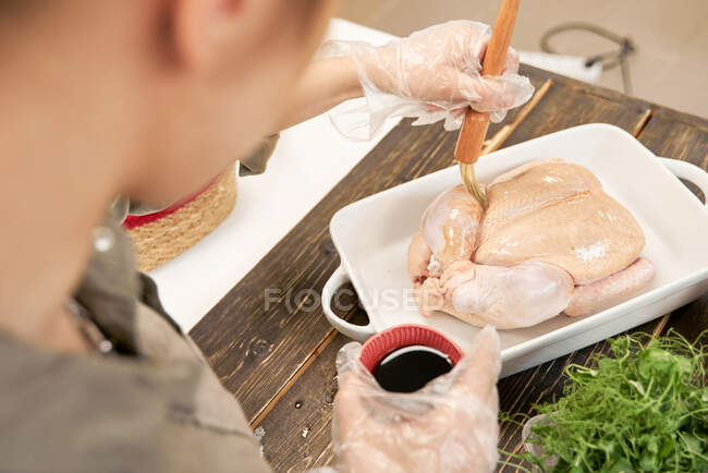 De cima da cultura fêmea anônima com escova de pastelaria untando aves cruas com molho de soja enquanto cozinha em casa — Fotografia de Stock