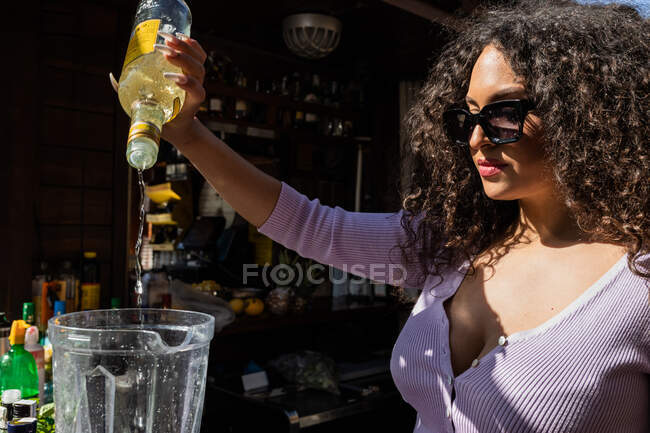 Contenido joven camarera étnica femenina en ropa casual añadiendo licor a la licuadora mientras mezcla bebidas en el soleado bar al aire libre - foto de stock