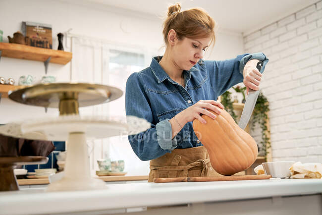 Jeune femme avec couteau tranchant coupant la courge crue sur la planche à découper tout en cuisinant dans la cuisine à la maison — Photo de stock