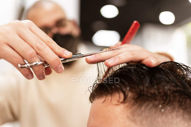 Male stylist trimming wet hair of crop anonymous client with scissors in hairdressing salon on blurred background — Foto stock