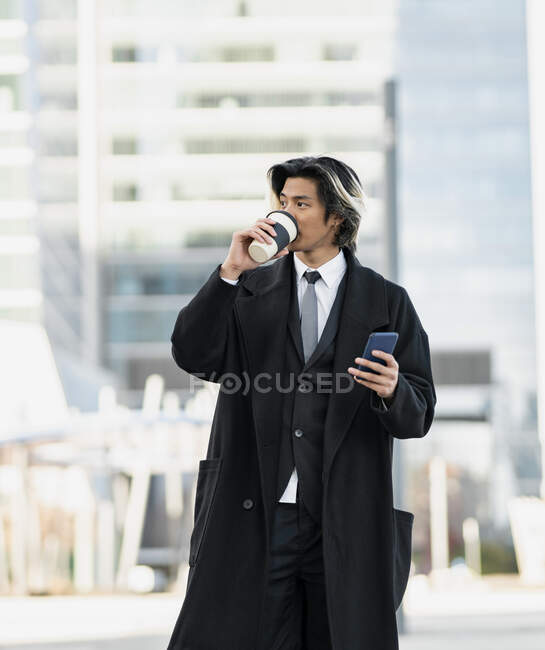 Молодой хорошо одетый этнический мужчина исполнительный с сотовым телефоном и горячим напитком, чтобы пойти в город глядя в сторону — стоковое фото