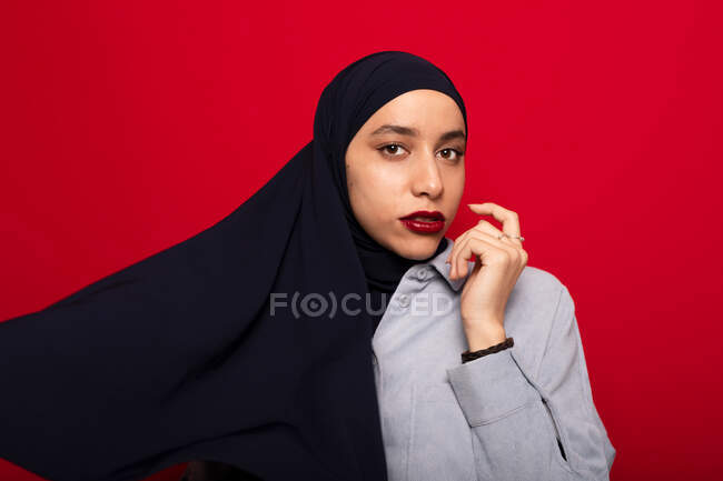 Jovem mulher islâmica atraente em camisa casual e hijab escuro em pé no fundo vermelho e olhando para a câmera — Fotografia de Stock