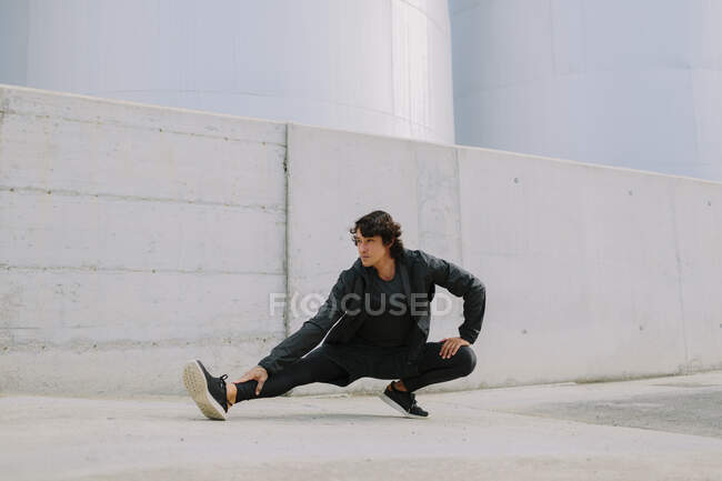 Homem de corpo inteiro em roupas esportivas pretas totais inclinadas com perna no chão enquanto se alonga no distrito industrial da cidade — Fotografia de Stock
