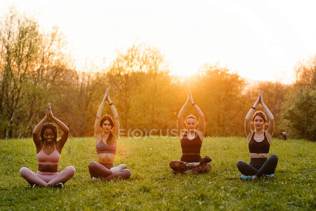 Compagnie de diverses femmes en vêtements de sport assis dans Lotus Poser avec les mains Namaste et pratiquer le yoga ensemble sur la pelouse dans le parc tout en regardant la caméra sur fond de ciel couchant — Photo de stock