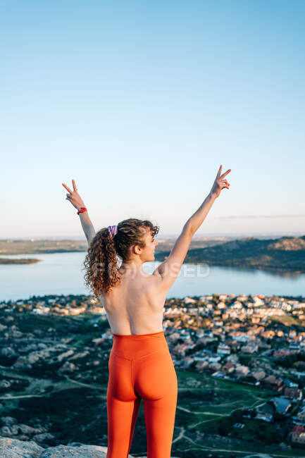 Back view of happy topless female traveler with curly hair in leggings smiling and showing two fingers gesture with raised arms standing on rocky cliff above coastal town — Stock Photo