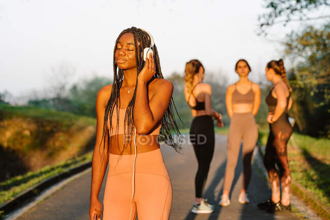 Cheerful African American female athlete listening to music in headphones while standing in park at sunset on background of company of blurred sportswomen — Stock Photo