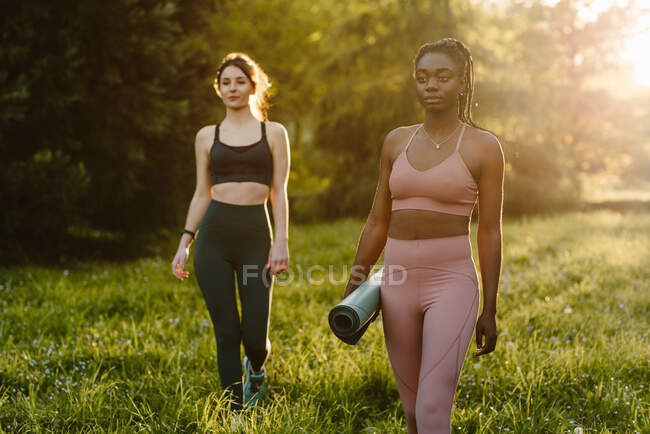 Confident fit multiracial female athletes walking with mat along lawn during training in summer park in evening — Stock Photo