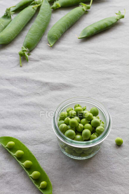 Desde arriba de un tazón pequeño de vidrio con guisantes verdes crujientes cerca de una pila de vainas de guisantes crujientes en la mesa en casa - foto de stock