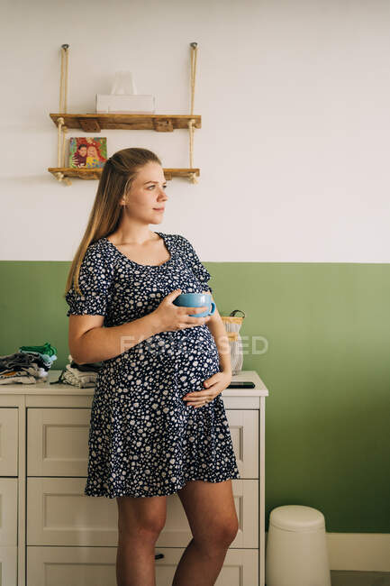 Молодая беременная женщина в декоративном платье с чашкой горячего напитка, глядя в сторону, гладя живот в комнате — стоковое фото