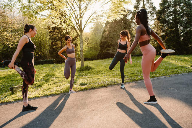 Young multiracial female athletes in sportswear stretching legs on asphalt footpath in town on sunny day — Stock Photo