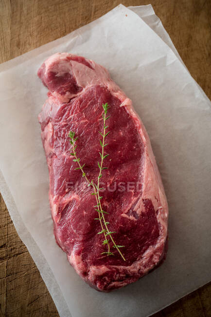 Vista aérea de la pieza de carne sin cocer con hojas de tomillo contra papel de hornear sobre fondo marrón - foto de stock