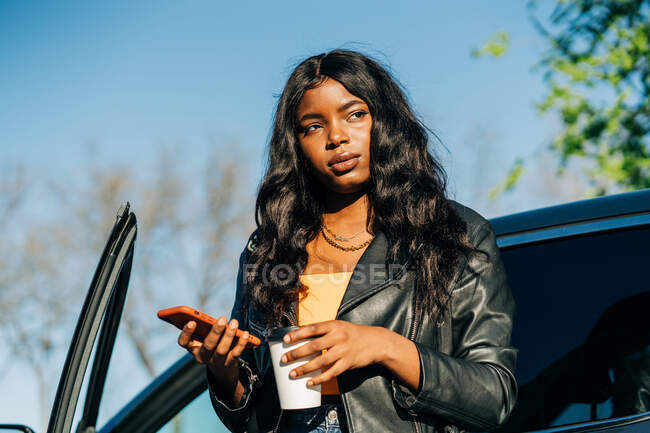 Mujer afroamericana segura con teléfono inteligente y bebida para llevar de pie cerca de un coche de lujo negro y mirando hacia otro lado - foto de stock