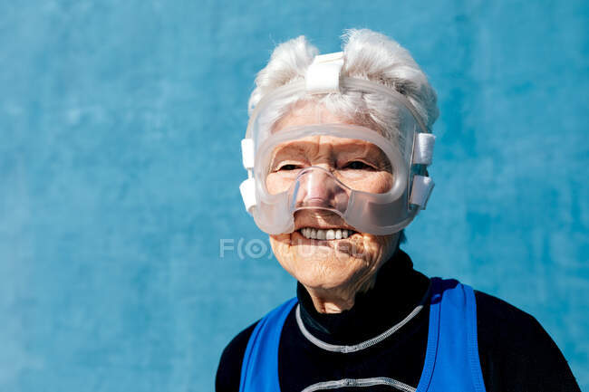Cheerful mature female with gray hair wearing protective boxing headgear against blue wall and looking away with smile — Stock Photo