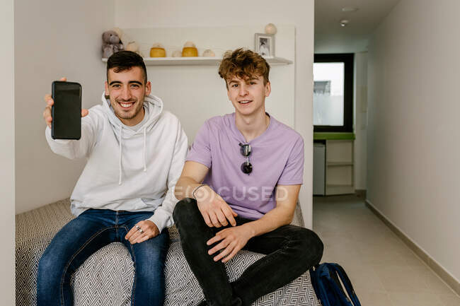 Cheerful young multiracial male friends in casual clothes sitting on bed and demonstrating mobile phone in modern apartment — Photo de stock