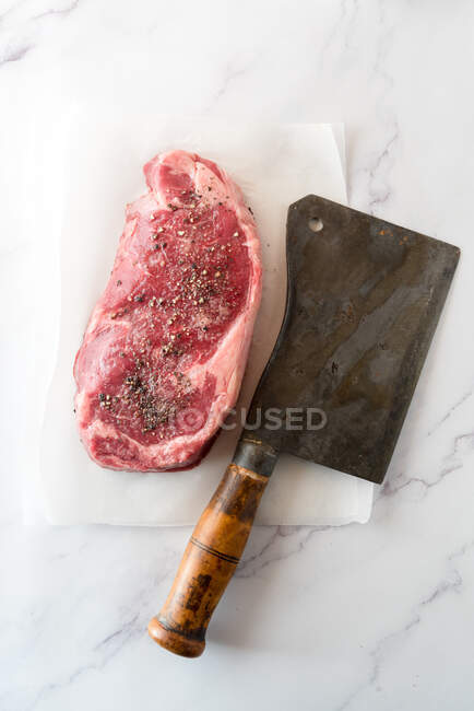 Overhead view of uncooked meat piece with black pepper against baking paper and hatchet knife on marble background — Stock Photo