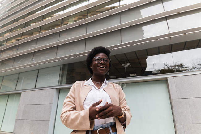 Low angle of delighted African American female entrepreneur with hair and in smart casual outfit standing against glass building in downtown and looking away — Stock Photo