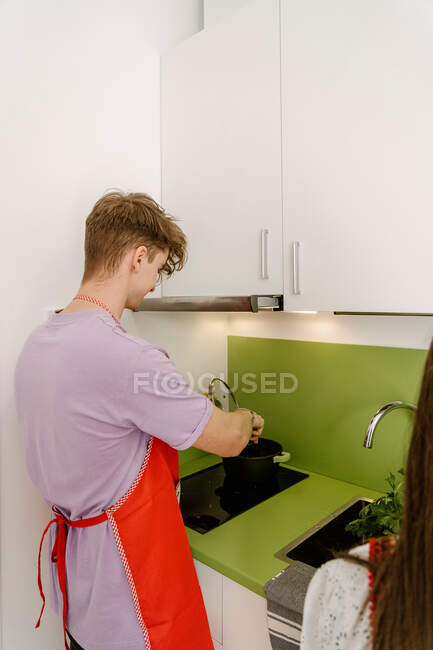 Side view of positive young male in casual clothes and apron stirring food in saucepan while preparing dinner in kitchen standing near crop unrecognizable girlfriend — Photo de stock