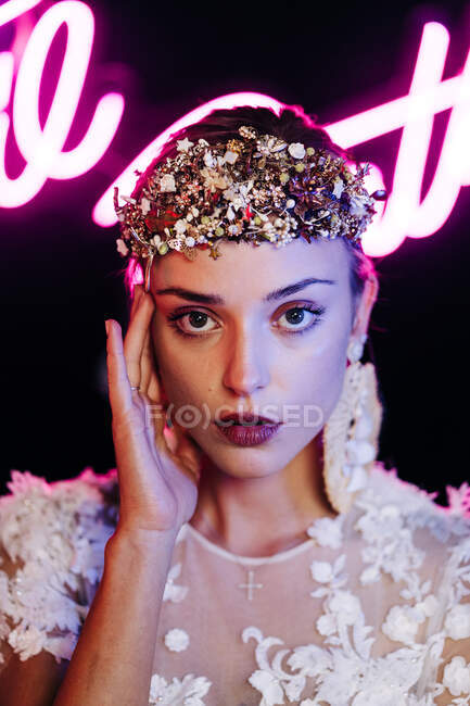 Charmante zarte junge Braut in weißem Spitzenkleid und luxuriösem Blumenkranz und Ohrringen blickt vor schwarzem Hintergrund mit Neonlicht in die Kamera — Stockfoto