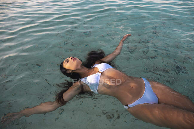 From above of young female in swimwear with outstretched arms and closed eyes lying in ocean with transparent water — Stock Photo