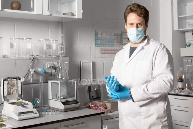 Chimico maschile in maschera sterile e guanti guardando la fotocamera contro l'analizzatore di umidità e l'equilibrio analitico nel laboratorio di cannabis — Foto stock