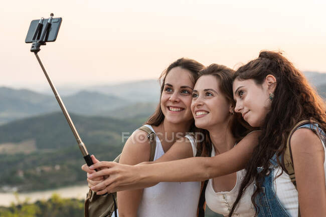 Путешествующие подруги с рюкзаками стоят на холме и делают самоснимок на смартфоне на фоне горного хребта летом — стоковое фото