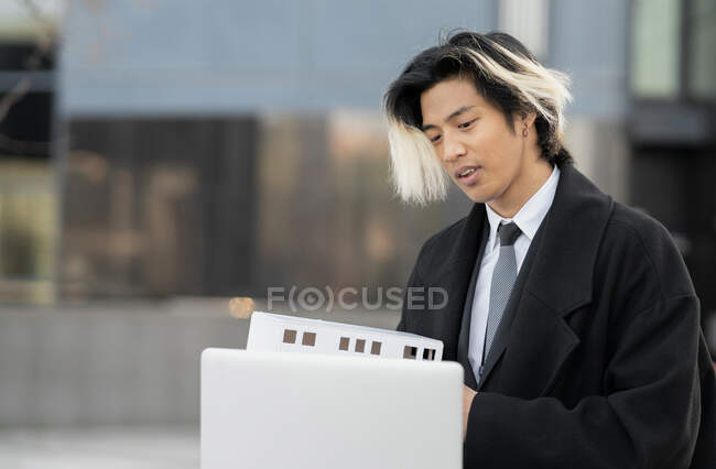 Joven empresario étnico masculino con maqueta de construcción hablando en videollamada contra netbook en la ciudad - foto de stock
