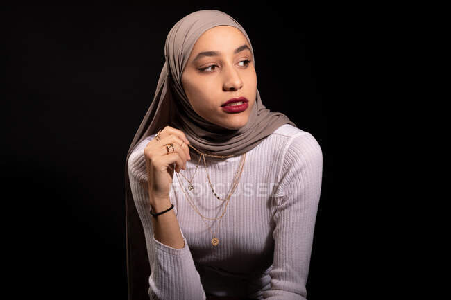 Модишь уверенную мусульманку в хиджабе, опираясь на стул и отводя взгляд в темную студию — стоковое фото
