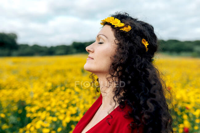 Vista lateral de la mujer de moda en vestido rojo y con corona de flores de pie con los ojos cerrados en el campo de flores con flores amarillas y rojas disfrutando de un cálido día de verano - foto de stock