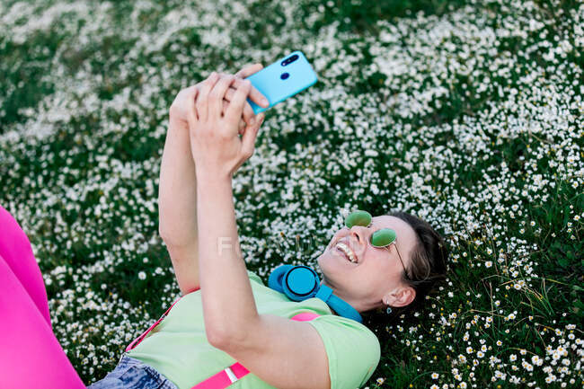 Side view joyful young female wearing bright outfit lying on lush grass with raised legs and browsing mobile phone in countryside — Stock Photo
