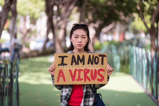 Етнічна жінка з картонним плакатом з написом Я не вірус, що протестує на вулиці міста і дивиться на камеру — стокове фото