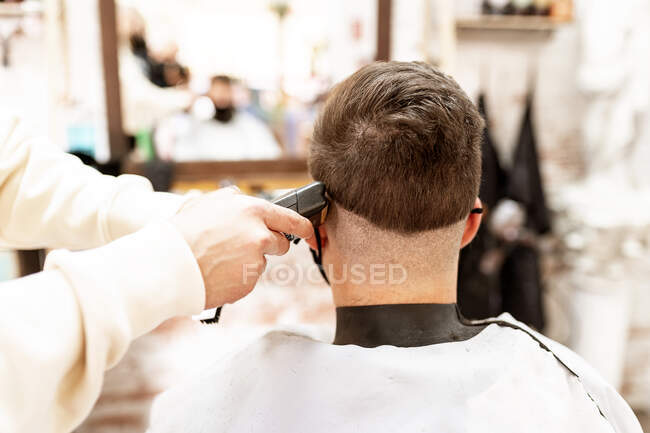 Crop anonymous male stylist with trimmer cutting hair of client in cape in barbershop — Stock Photo