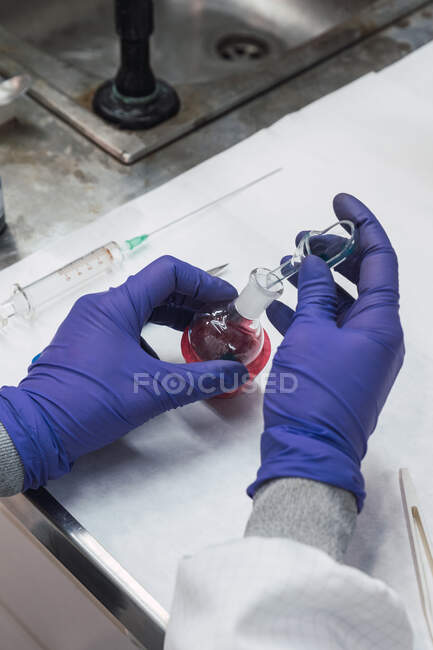 Сверху анонимный химик в латексных перчатках, наполняющий стеклянный пипетку химической жидкостью из кипящей фляжки во время проведения теста в лаборатории — стоковое фото