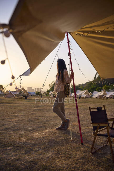 Étnica mujer asiática tener un tiempo relajante de pie mirando hacia otro lado en el área de camping durante la puesta del sol - foto de stock