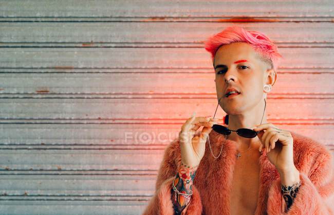Jeune gay en tenue élégante avec tatouage, cheveux roses et manucure avec veste moelleuse tenant des lunettes de soleil tout en regardant la caméra — Photo de stock