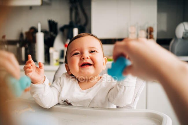 Cute laughing baby in white shirt sitting in baby feeding chair in modern kitchen — Stock Photo