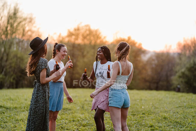 Anonimo migliori amiche multirazziali con bottiglie di bevanda alcolica guardarsi mentre si parla sul prato in serata — Foto stock