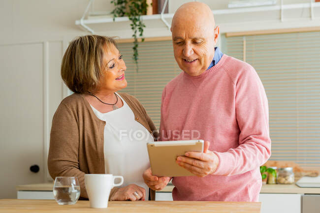 Amar pareja de mediana edad de pie en la mesa en la cocina y navegar por Internet en la tableta - foto de stock