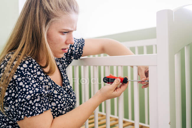 Side view woman with screwdriver leaning forward while mounting crib at home on sunny day — Stock Photo