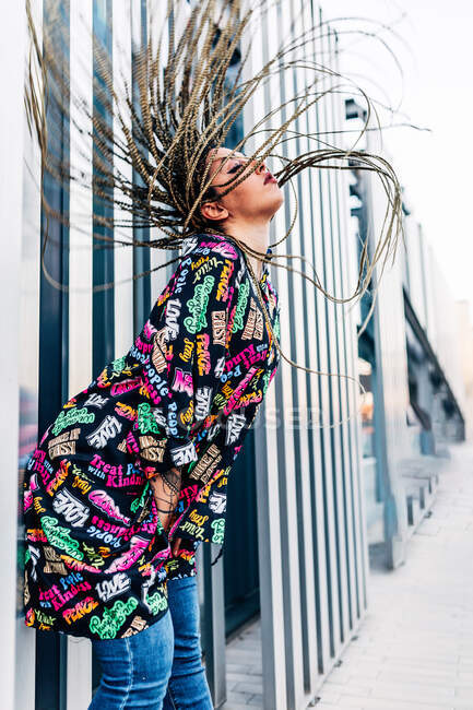 Seitenansicht einer Millennial-Frau mit langen Afro-Zöpfen, die sich vorwärts gegen die Wand auf dem städtischen Gehweg lehnt — Stockfoto