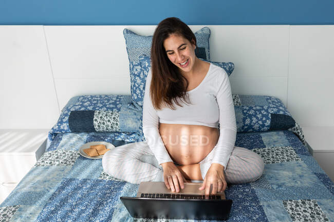 Dall'alto di una donna incinta sorridente che parla in videochiamata su netbook mentre è seduta su un comodo letto a casa — Foto stock