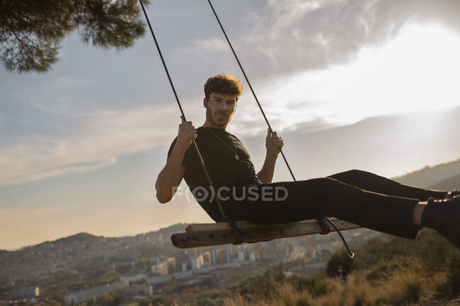 Bearded male in black apparel looking at camera while sitting on swing against mountains in town at sundown — Stock Photo