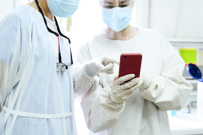 Female surgeons in medical cloth caps surfing internet on cellphone against desktop computer in light hospital — Stock Photo