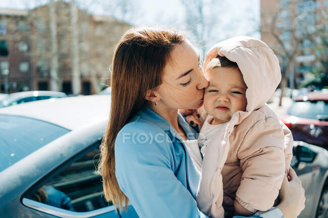 Aimant jeune mère tenant sur les mains et embrassant adorable petite joue de bébé tout en se tenant debout sur la rue ensoleillée du printemps — Photo de stock