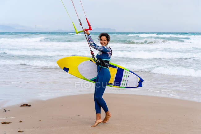 Alegre kiter femenino en traje de neopreno sosteniendo la barra de control mientras mira la cámara en la orilla del océano arenoso - foto de stock