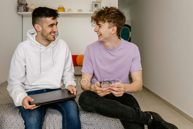 Fröhliche junge, unterschiedliche männliche beste Freunde lachen und schauen einander an, während sie auf einem bequemen Bett mit Laptop und Smartphone sitzen — Stockfoto