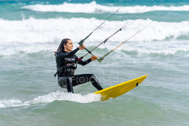 Atleta femenina activa en el kiteboard sosteniendo la barra de control mientras practica kitesurf y mira hacia otro lado en el océano espumoso - foto de stock