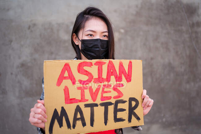 Mujer étnica enmascarada y con cartel de cartón con inscripción Asian Lives Matter protestando en la calle de la ciudad y mirando hacia otro lado - foto de stock