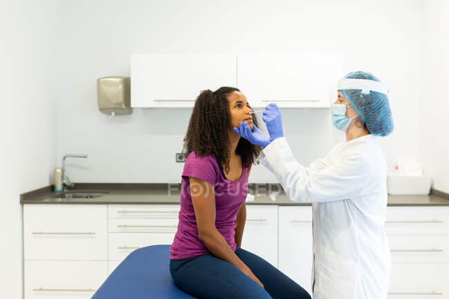 Especialista médica femenina en uniforme protector, guantes de látex y mascarilla para la cara haciendo una prueba de hisopo nasal con brote de algodón en una paciente afroamericana en una clínica durante el brote de coronavirus - foto de stock