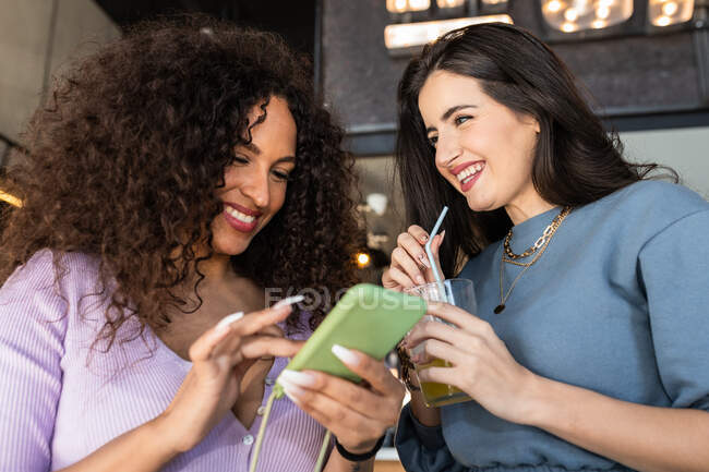 Улыбающиеся юные подруги в повседневной одежде, просматривающие мобильные телефоны, выпивая газировку в ресторане — стоковое фото