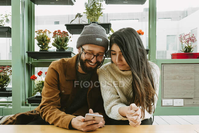 Pareja de contenido sentada en la mesa y navegando juntos por el teléfono móvil mientras se enfría en la cafetería con una variedad de flores en maceta - foto de stock
